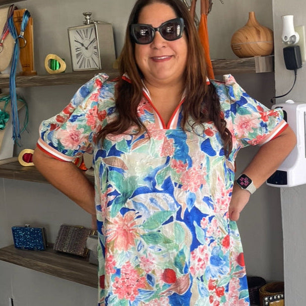 Woman wearing a colorful floral dress in an indoor setting with shelves and decor.
